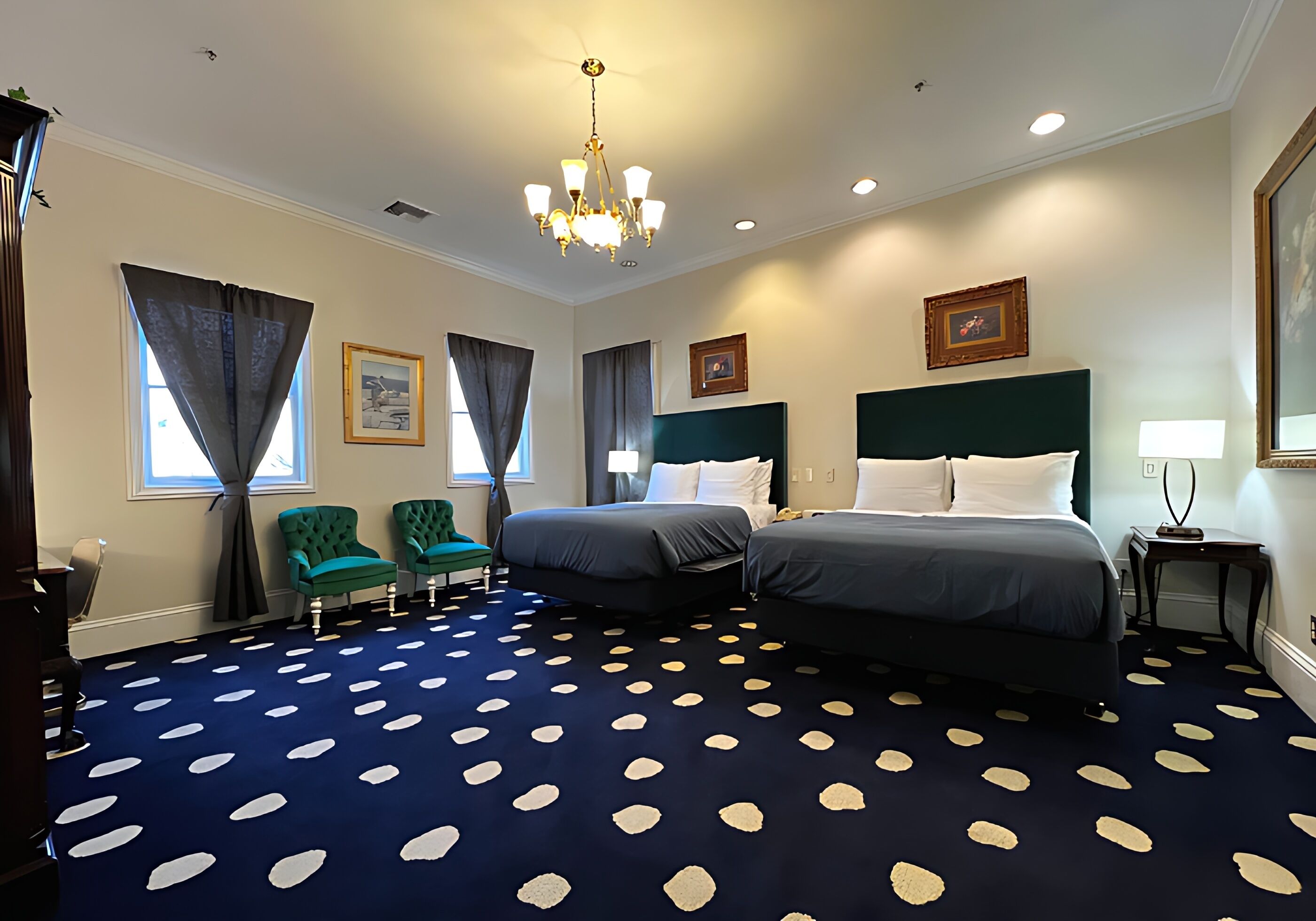 Une chambre d’hôtel avec deux lits, un lustre, une moquette bleue à motifs blancs et des fauteuils verts.