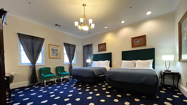 Een hotelkamer met twee bedden, een kroonluchter, een blauw tapijt met witte patronen en groene fauteuils.