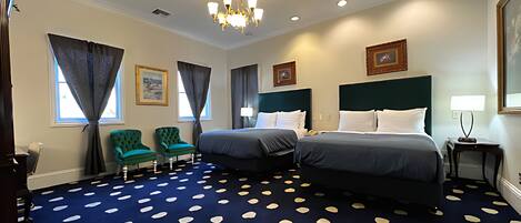 Habitación de hotel con dos camas, una araña y una alfombra azul con diseños blancos, además de sillones verdes.