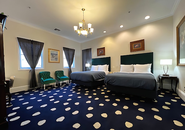 Habitación de hotel con dos camas, una araña y una alfombra azul con diseños blancos, además de sillones verdes.