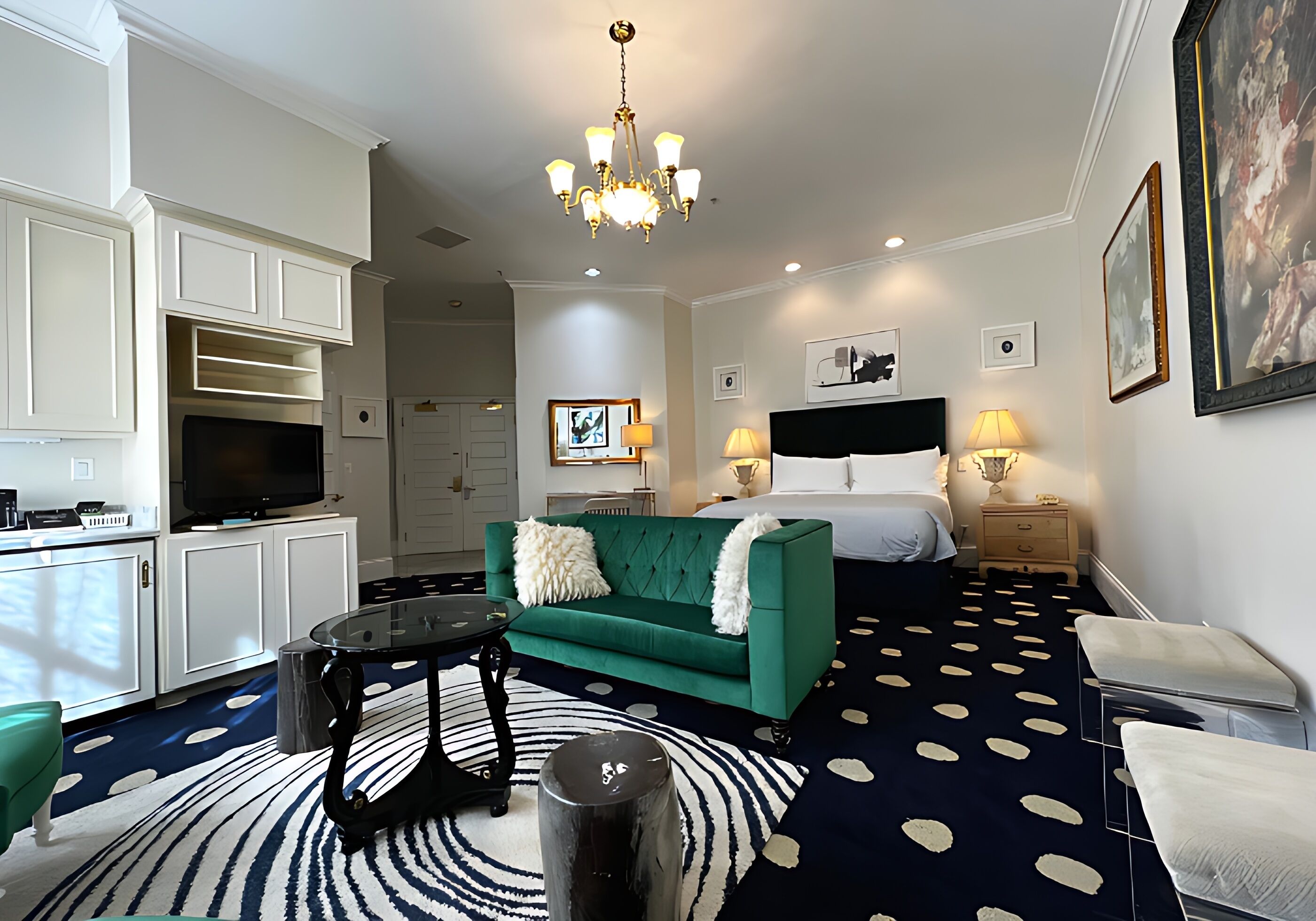 Ein Hotelzimmer mit einem grünen Sofa, einem runden Couchtisch, einem Fernseher, einem Bett mit weißer Bettwäsche und einer Kristallleuchter.