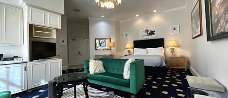 Ein Hotelzimmer mit einem grünen Sofa, einem runden Couchtisch, einem Fernseher, einem Bett mit weißer Bettwäsche und einer Kristallleuchter.