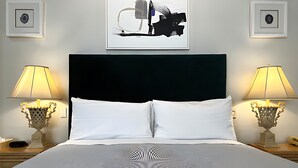 Et hotelværelse med en stor seng, to sengeborde med lamper, en sort sengegavl og abstrakt kunst på væggen.