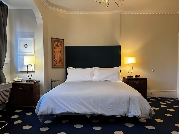 Quarto de hotel com uma cama grande, duas mesas de cabeceira com luminárias, uma obra de arte emoldurada na parede e um tapete estampado.