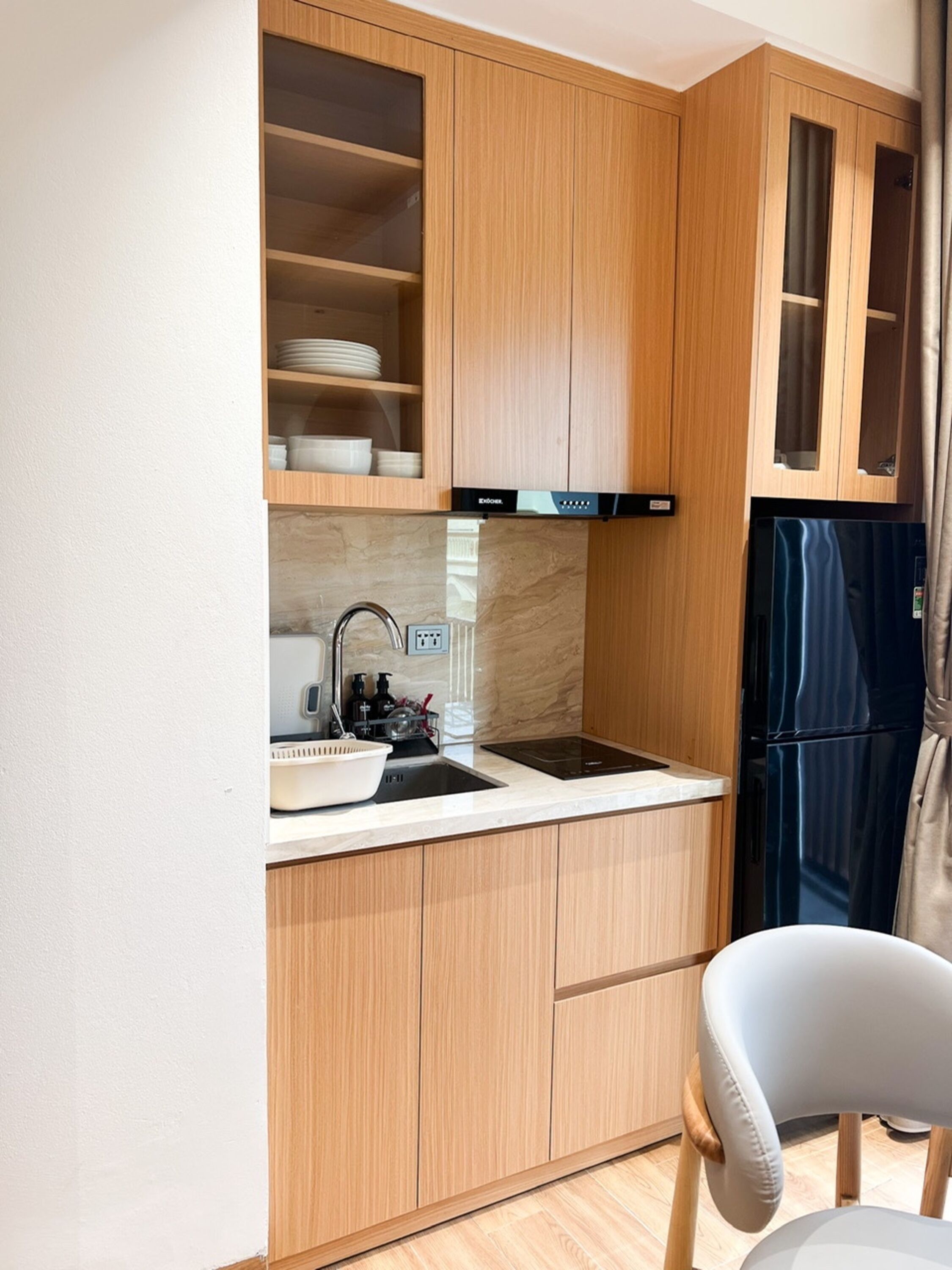 A compact kitchen with wooden cabinets, a sink, a black refrigerator, and a chair.