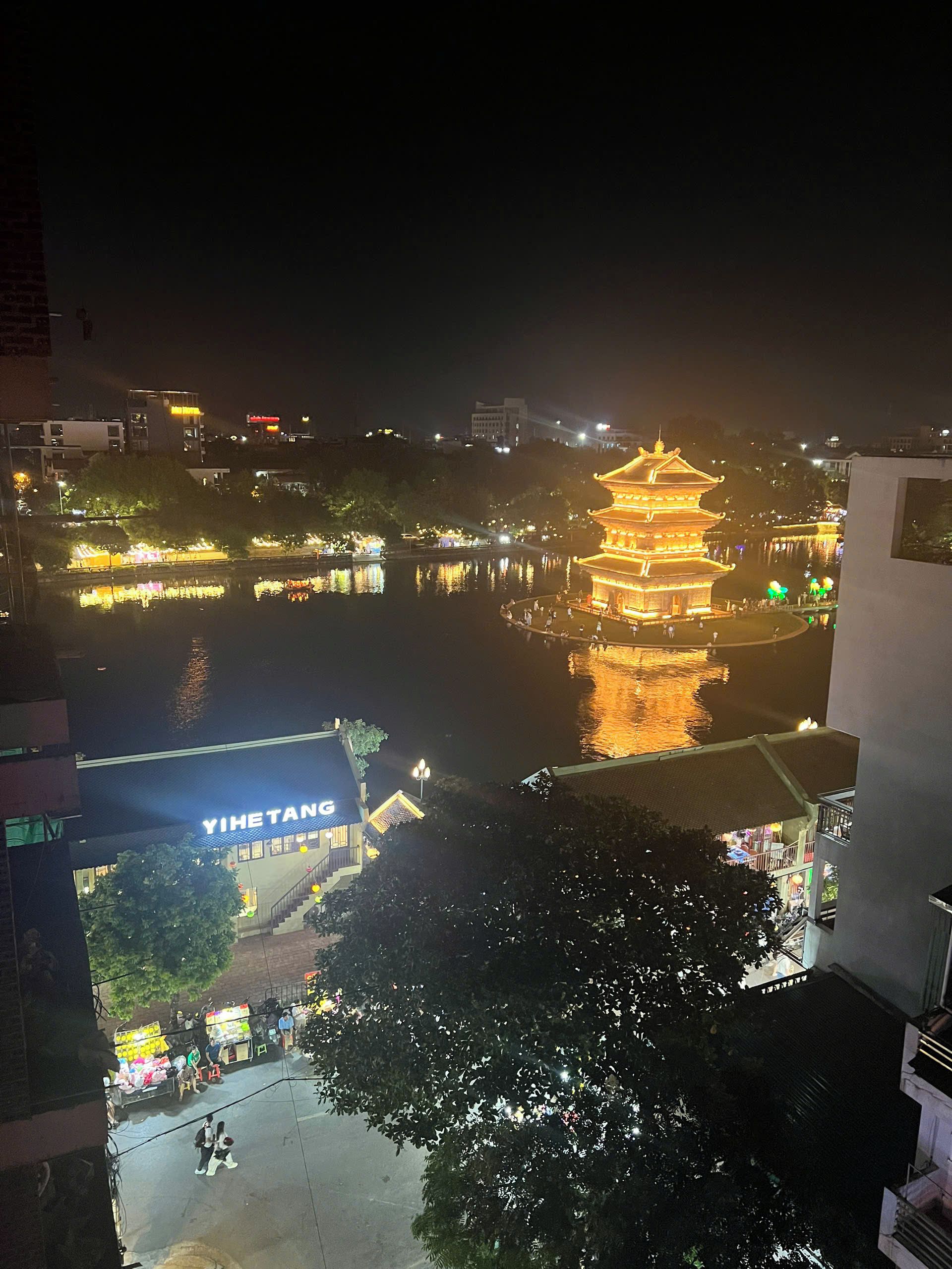 A traditional pagoda with a reflection in the water, a building with a sign 'YIHTANG', and a tree in the foreground.