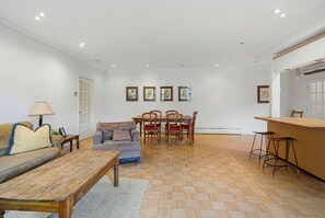 A living room with a sofa, a coffee table, a dining table with chairs, and a kitchen area with stools.
