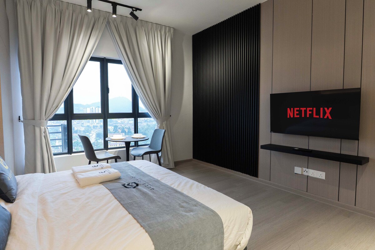 A hotel room with a bed, a TV displaying 'NETFLIX,' a dining table with chairs, and a view of a cityscape through large windows.