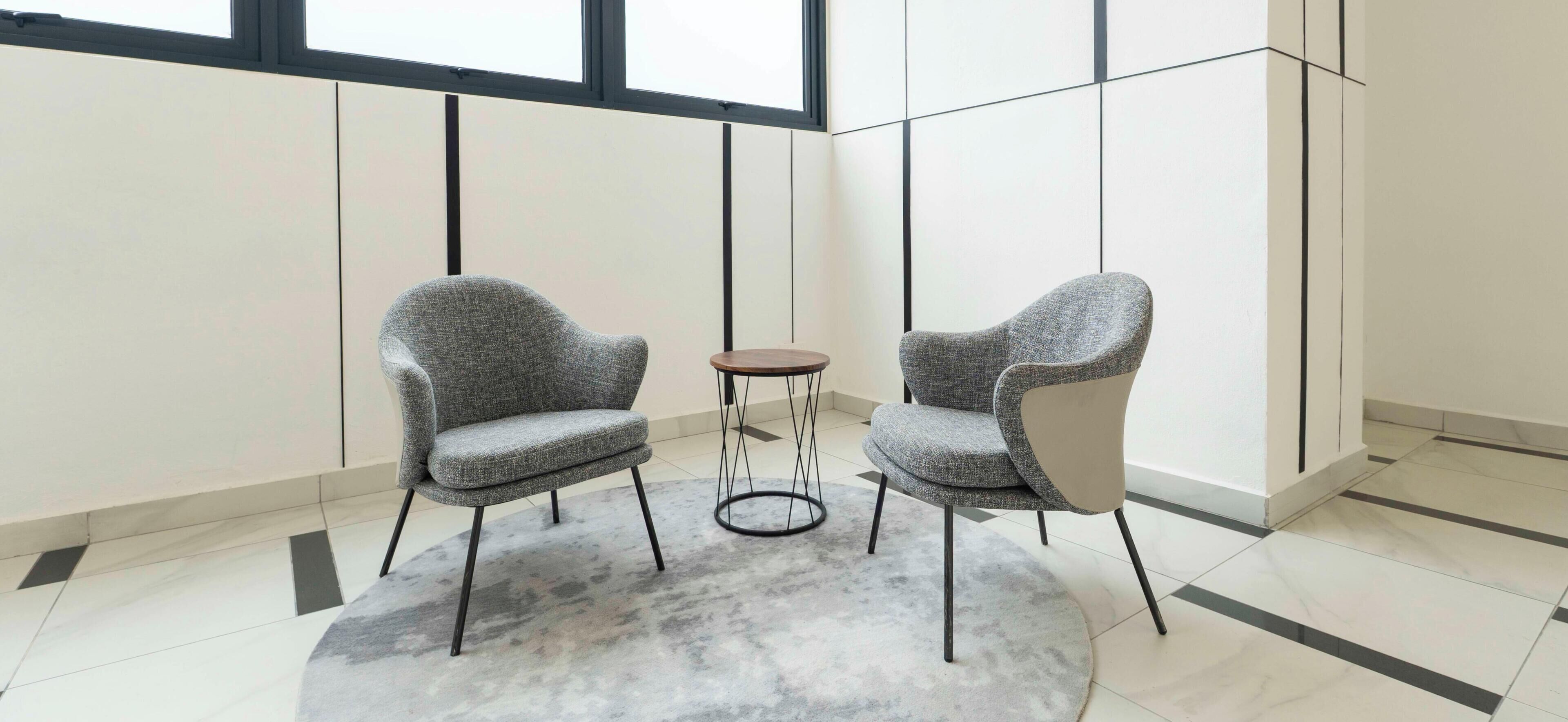 A modern room with two grey upholstered chairs, a small round table, and a grey carpet.