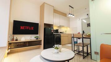 A modern living room with a flat-screen TV, a dining area with a table and chairs, a kitchen with appliances, and a small plant on the table.