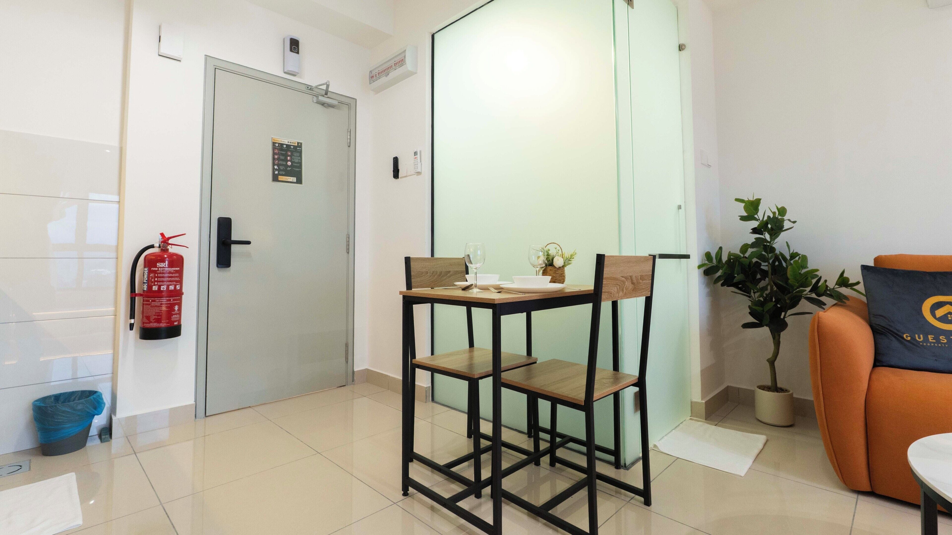 Habitación de hotel con puerta de vidrio, una mesita con sillas, extintor, planta en maceta y un cojín con un logotipo.