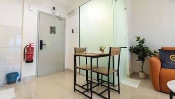 Habitación de hotel con puerta de vidrio, una mesita con sillas, extintor, planta en maceta y un cojín con un logotipo.