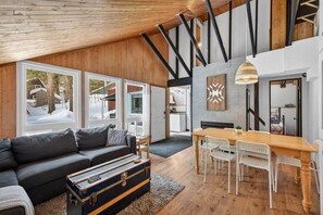 A modern interior with a wooden ceiling, a black sofa, a dining table with white chairs, and large windows.