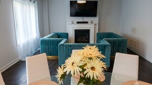 A living room with teal sofas, a white fireplace, a flat-screen TV, and a yellow vase with flowers.