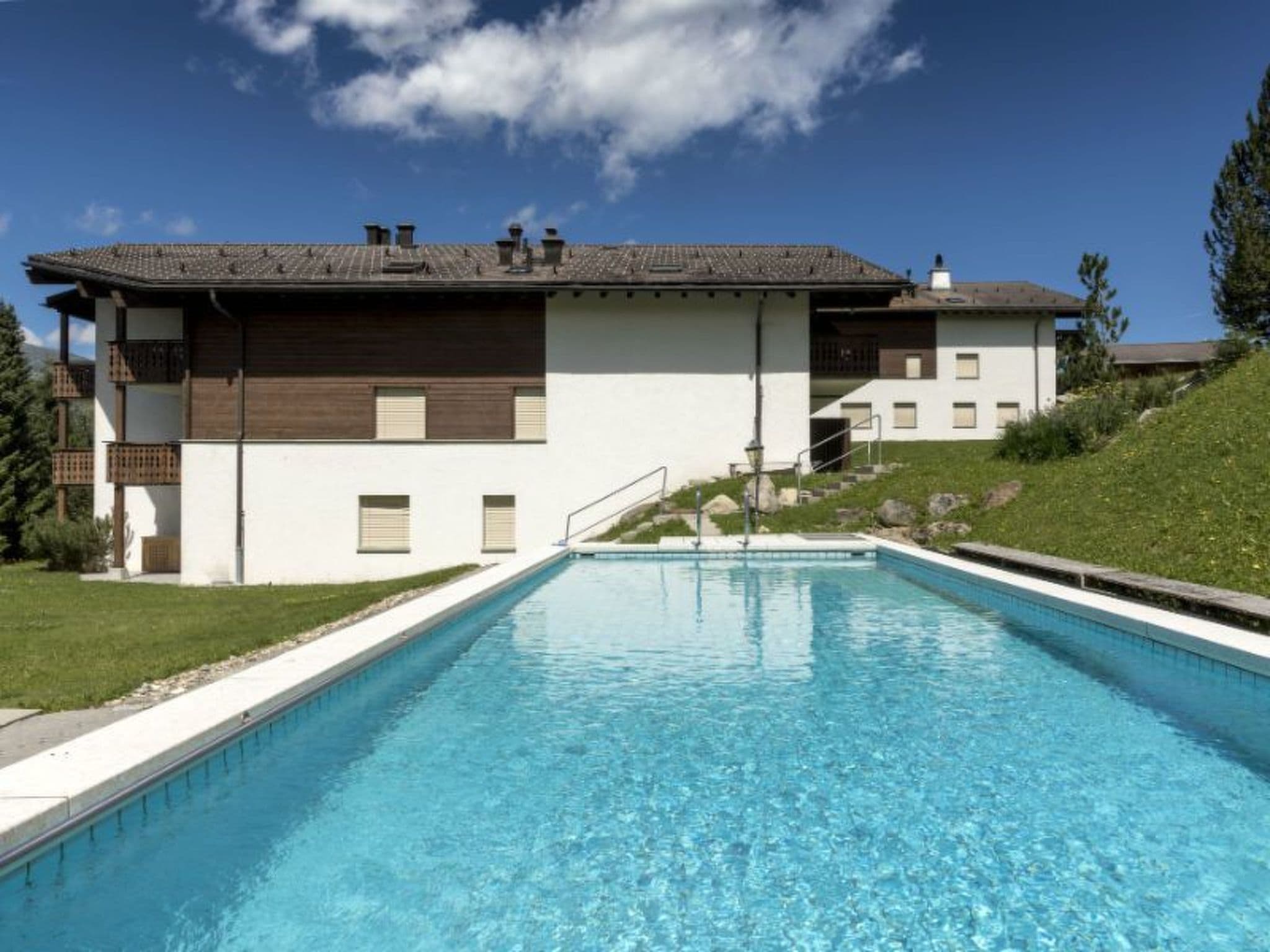 A swimming pool with a clear blue water surface, surrounded by a white building with a brown roof and a grassy area.