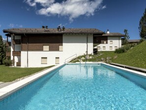 A swimming pool with a clear blue water surface, surrounded by a white building with a brown roof and a grassy area. - Gadastatt, Obersaxen-Valata, (Surcuolm), 38041B by Interhome (Obersaxen Surcuolm)