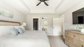 Une chambre à coucher comprenant un lit, une commode, un téléviseur, un ventilateur de plafond et une porte donnant sur une autre pièce.