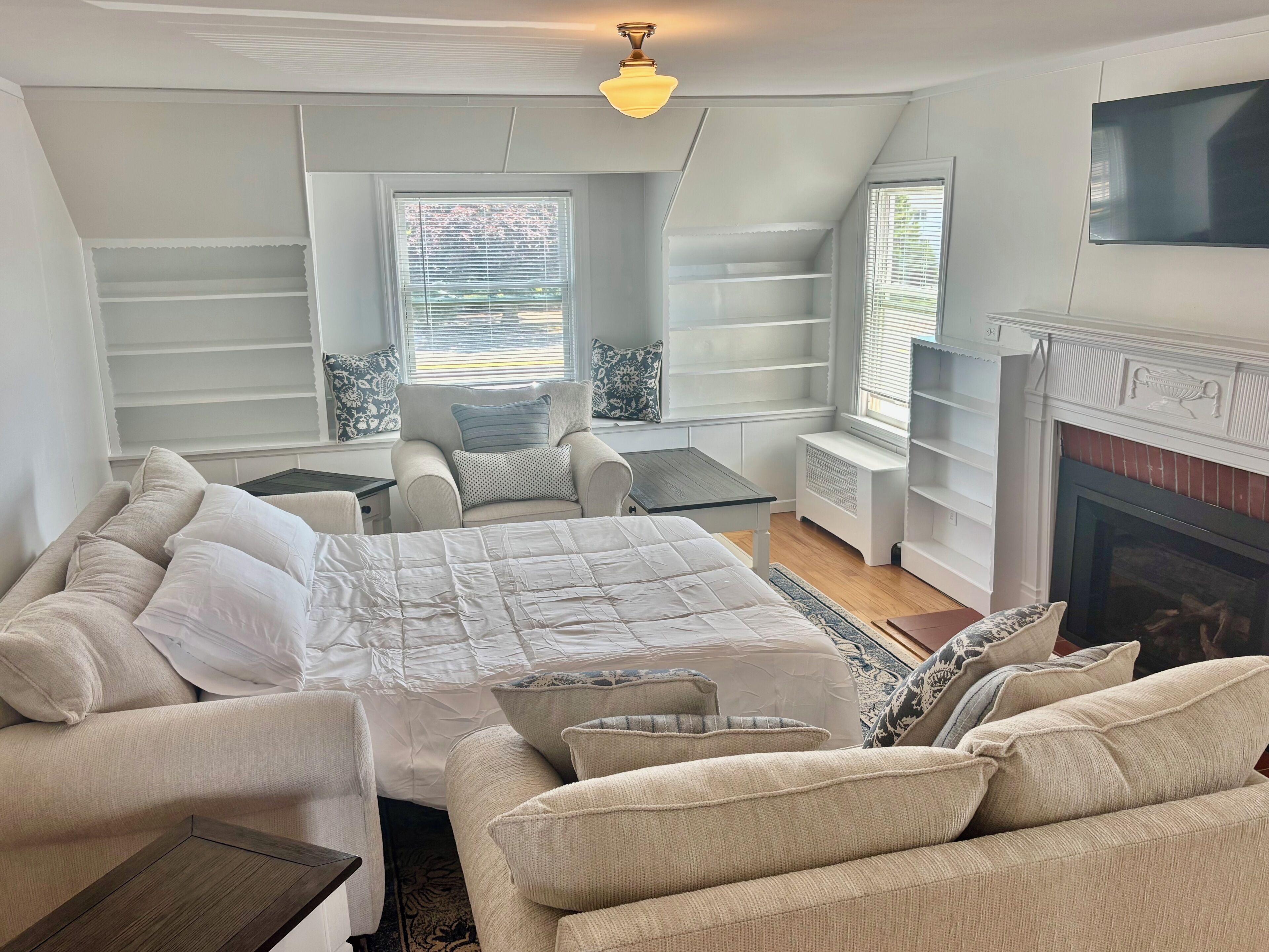 A bedroom with a bed, two armchairs, a small table, a fireplace, and built-in shelving.