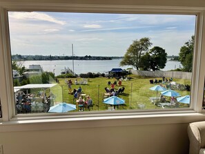 A view through a window of a backyard with a grassy lawn, outdoor seating, and a body of water in the background. - Year round 1-bedroom coastal LOFT, sleeps 6, in charming Portsmouth RI (Portsmouth)