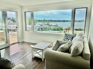 A room with a large window overlooking a body of water, a wooden floor, and a sofa with cushions. - Year round 1-bedroom coastal LOFT, sleeps 6, in charming Portsmouth RI (Portsmouth)