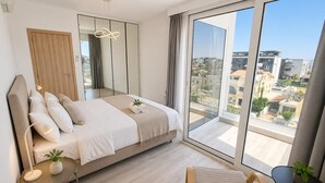 Een moderne slaapkamer met een bed, een stoel, een klein tafeltje met een plant, een spiegel en een groot raam met uitzicht op de stad.