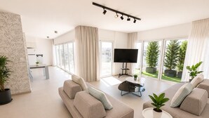 Een moderne woonkamer met een bank, een salontafel en een televisie. Er zijn grote ramen en een balkon met planten.