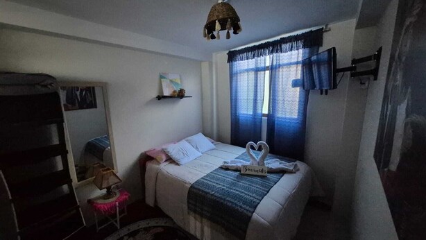 A bedroom with a bed, a bedside table, a shelf with a picture, and a window with curtains. - Charming 1-bedroom apartment in the heart of Cusco (Cusco)