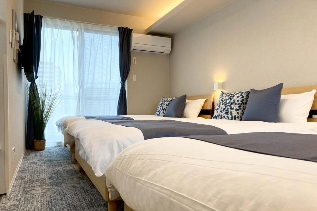 Ein Hotelzimmer mit drei Betten, einem großen Fenster mit Vorhängen, einer Klimaanlage und einem Blumentopf.