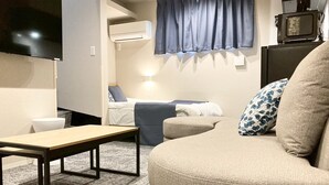 A living room with a sofa, a table, a TV, an air conditioning unit, and a bed with a blue curtain. - Koti Sopo Universal Bay 5 by Liaison (Osaka)