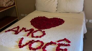 Uma cama com colcha e travesseiros brancos, um coração feito de pétalas de rosa com a frase 'I love you' escrita em pétalas vermelhas, e uma mesa de cabeceira com um travesseiro floral.