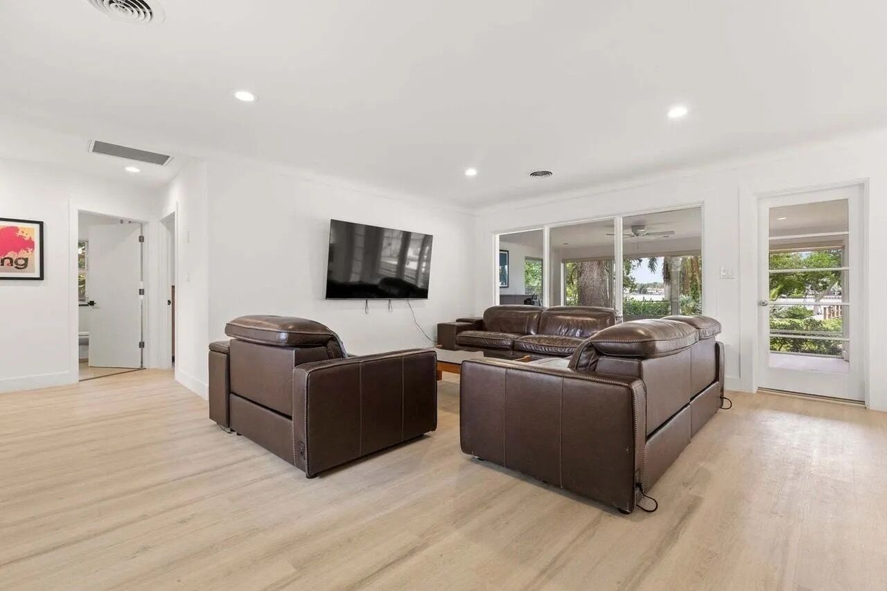 A spacious living room with a brown leather sofa set, a flat-screen TV mounted on the wall, and a large sliding glass door leading to a balcony.