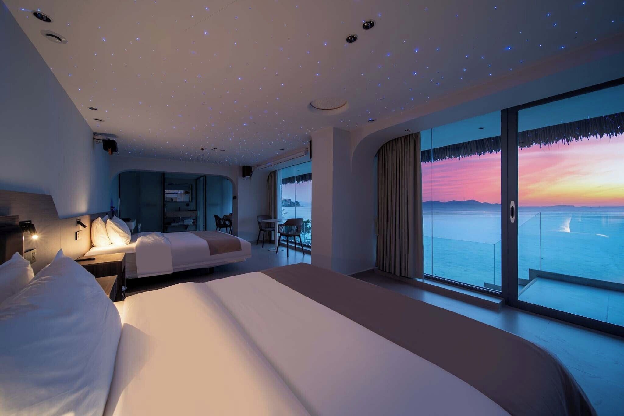 Una habitación de hotel moderna con una cama grande, una zona de estar y un balcón con vistas al mar al atardecer.