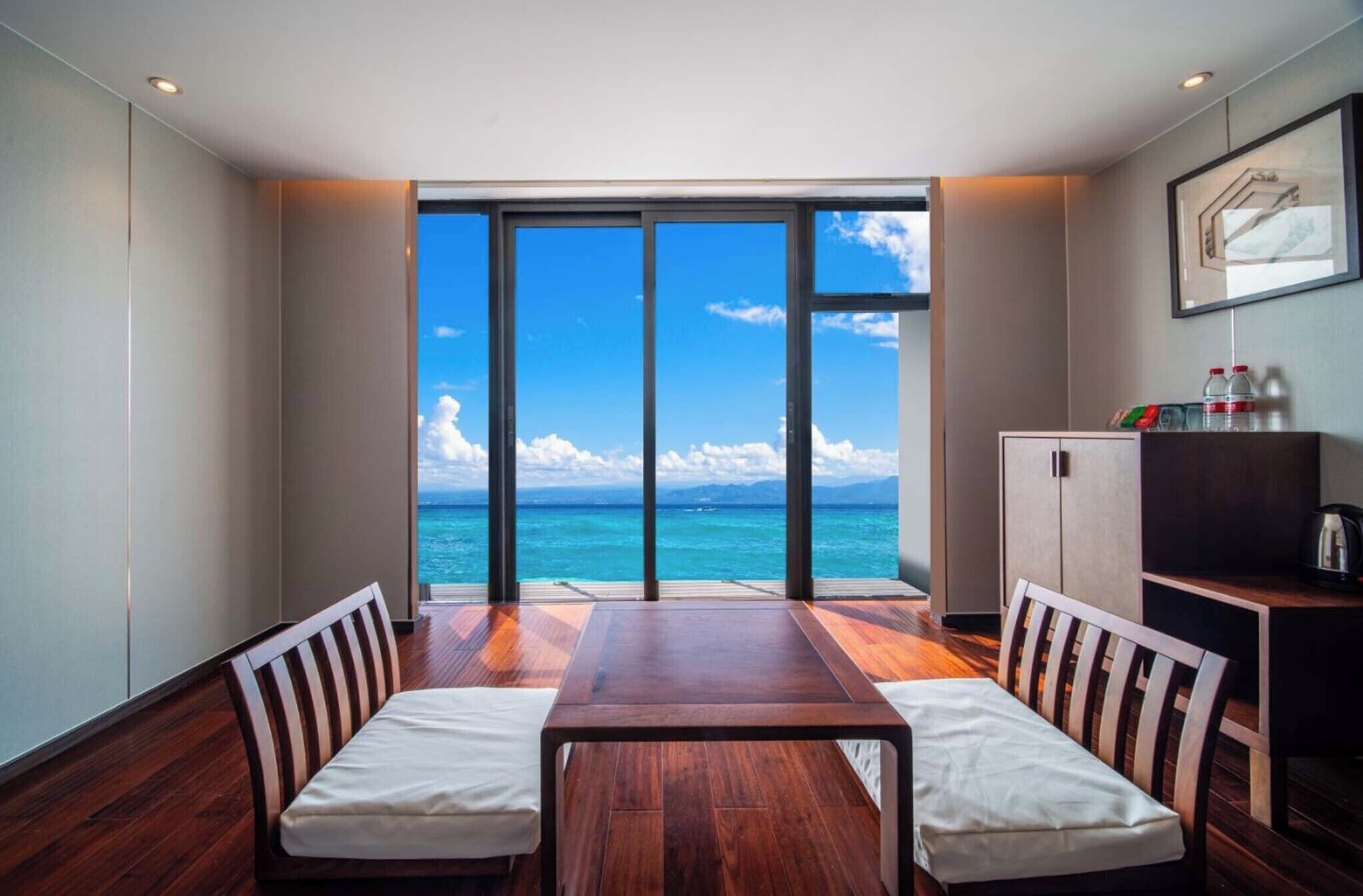 A room with a large sliding glass door leading to a balcony with a view of the ocean. There is a wooden table with two chairs and a cabinet with bottles on top.