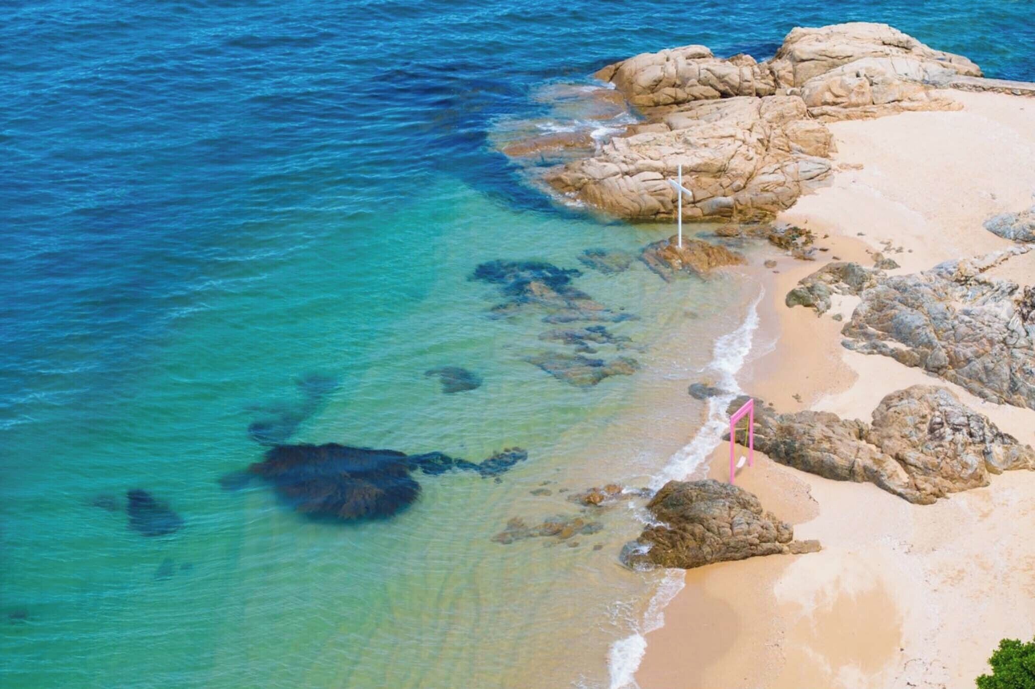 Una playa costera con aguas cristalinas, formaciones rocosas e una bandera en un asta.