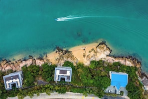 Vista aérea de una propiedad costera con playa de arena, costa rocosa y varios edificios de estilos arquitectónicos diferentes.