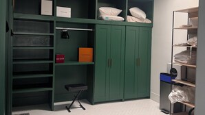 Um quarto com um guarda-roupa verde, prateleiras com caixas identificadas com 'GUCCI' e 'LOUIS VUITTON', uma pequena mesa e um chão com padrão de espinha de peixe.