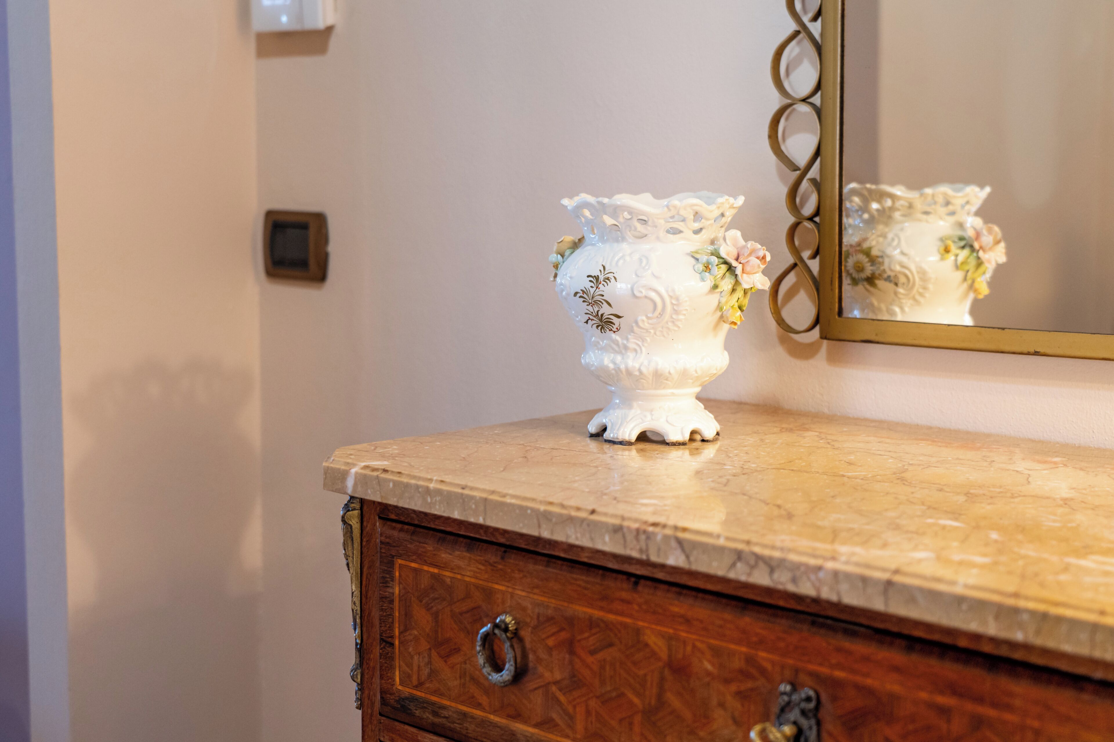 A white ceramic vase with floral designs on a marble countertop, a wooden dresser with a mirror, and a wall switch.