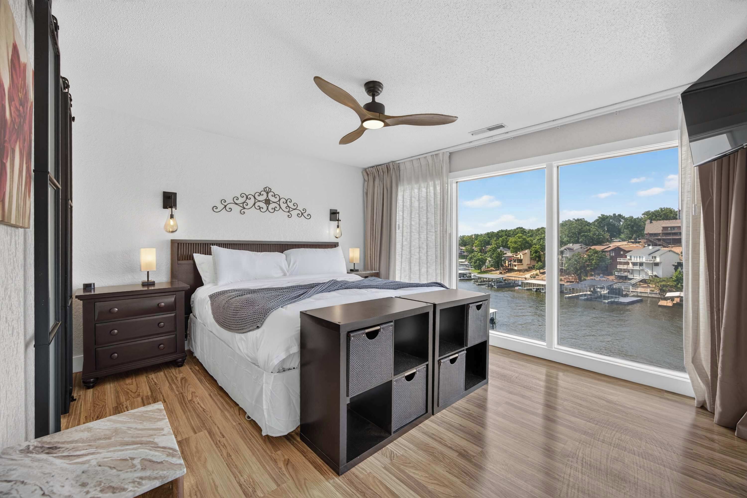 Une chambre à coucher comprenant un lit, des tables de chevet, un ventilateur de plafond, une grande fenêtre donnant sur une rivière et des bâtiments, ainsi qu'un plancher en bois.