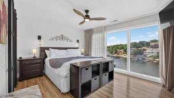 Une chambre à coucher comprenant un lit, des tables de chevet, un ventilateur de plafond, une grande fenêtre donnant sur une rivière et des bâtiments, ainsi qu'un plancher en bois.