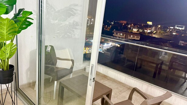 Un balcon avec une porte-fenêtre, un pot de fleurs, une chaise, une petite table et une vue sur le paysage urbain illuminé la nuit.