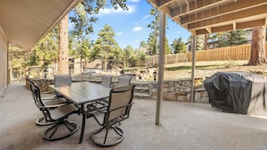 Un patio exterior con mesa y sillas, una zona cubierta y vista a un jardín cercado con árboles.