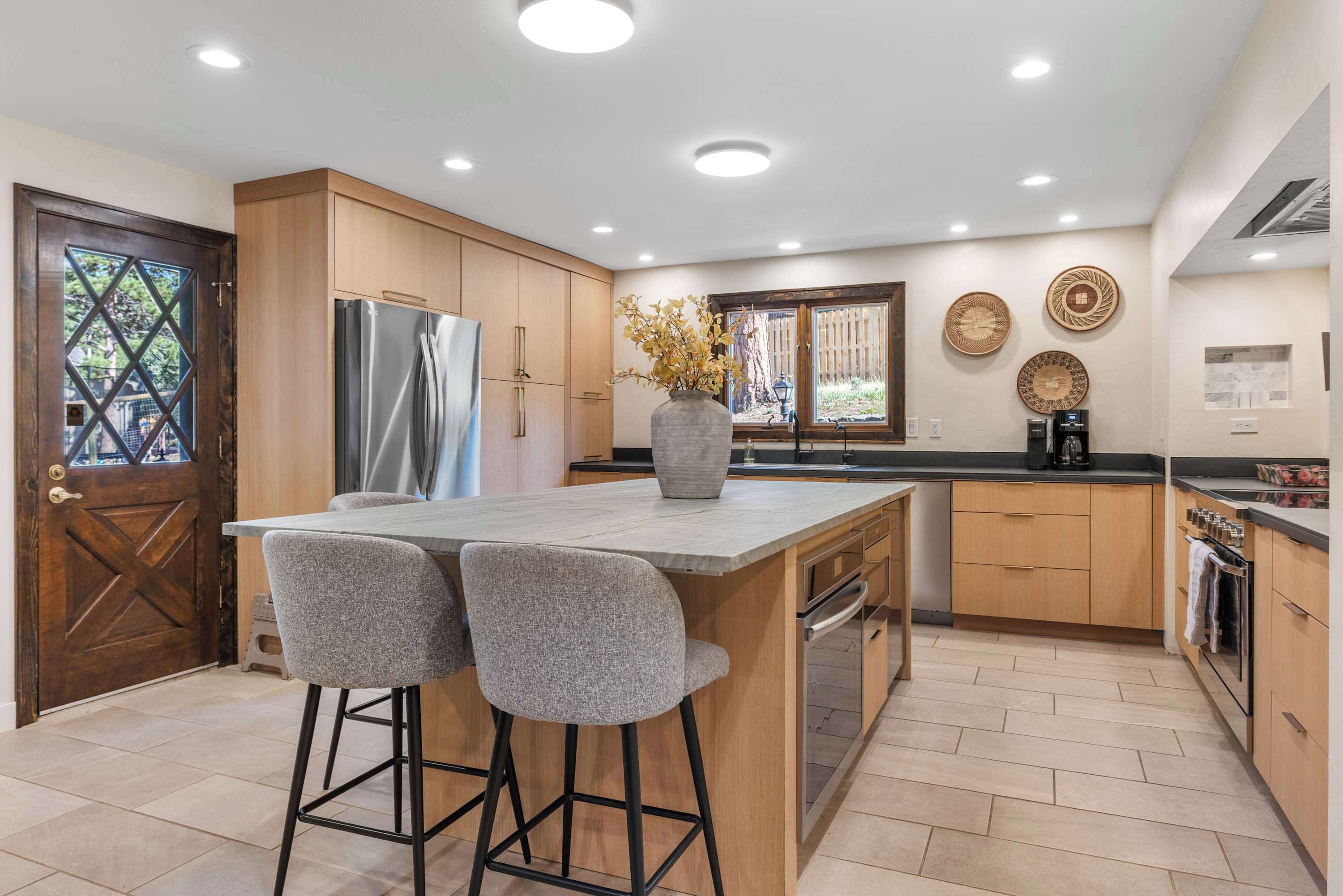 A modern kitchen with a central island, bar stools, wooden cabinets, and a large window.