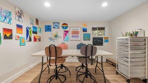 Un taller de arte con una mesa blanca, dos sillas y una pared llena de diversas obras.