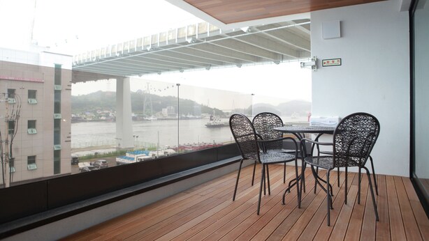 A balcony with a table and chairs, overlooking a river with boats and buildings in the background. - GITE Hotel Yeosu (Yeosu)