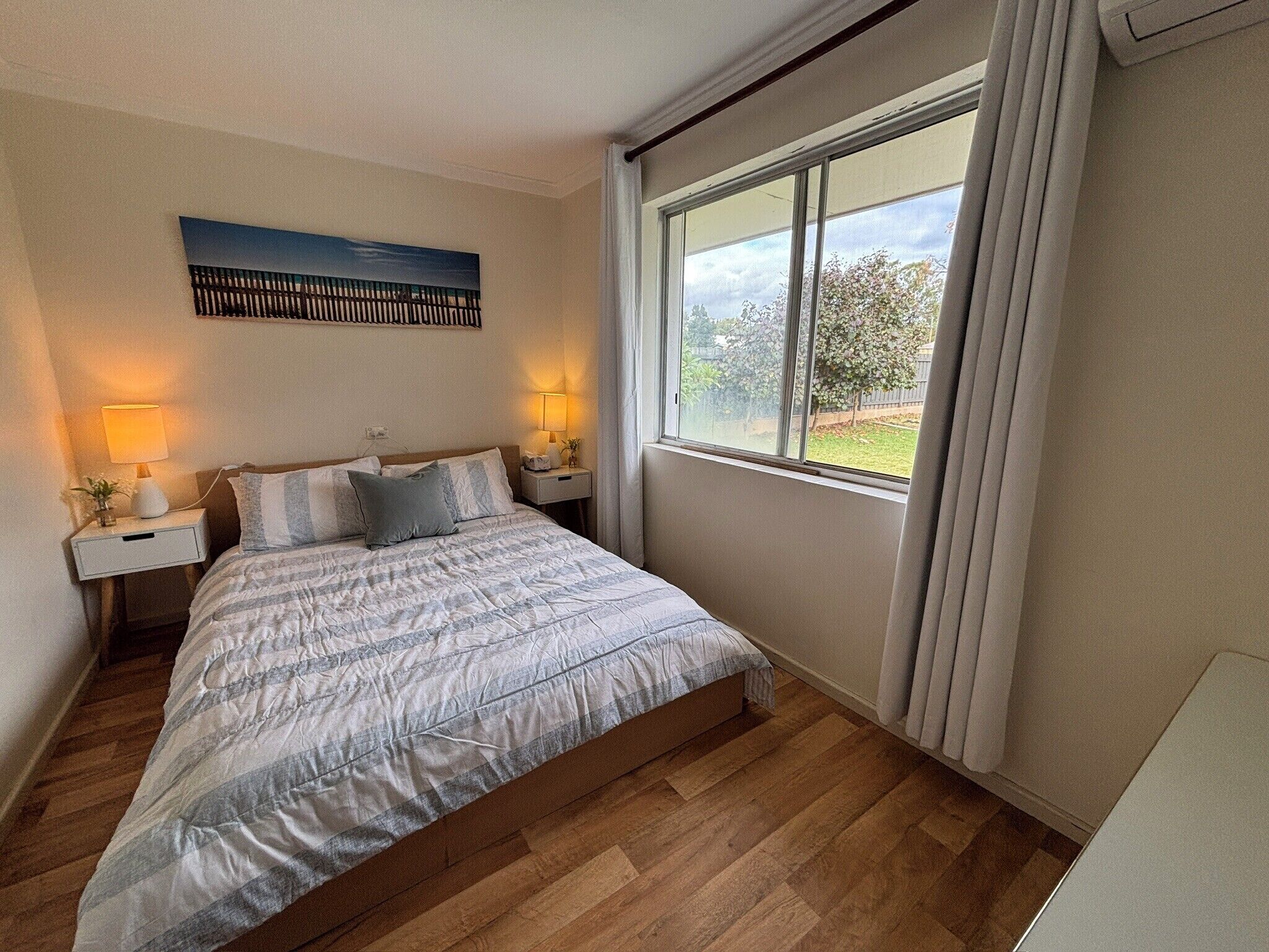 A bedroom with a large bed, two bedside tables with lamps, a window with curtains, and a painting on the wall.