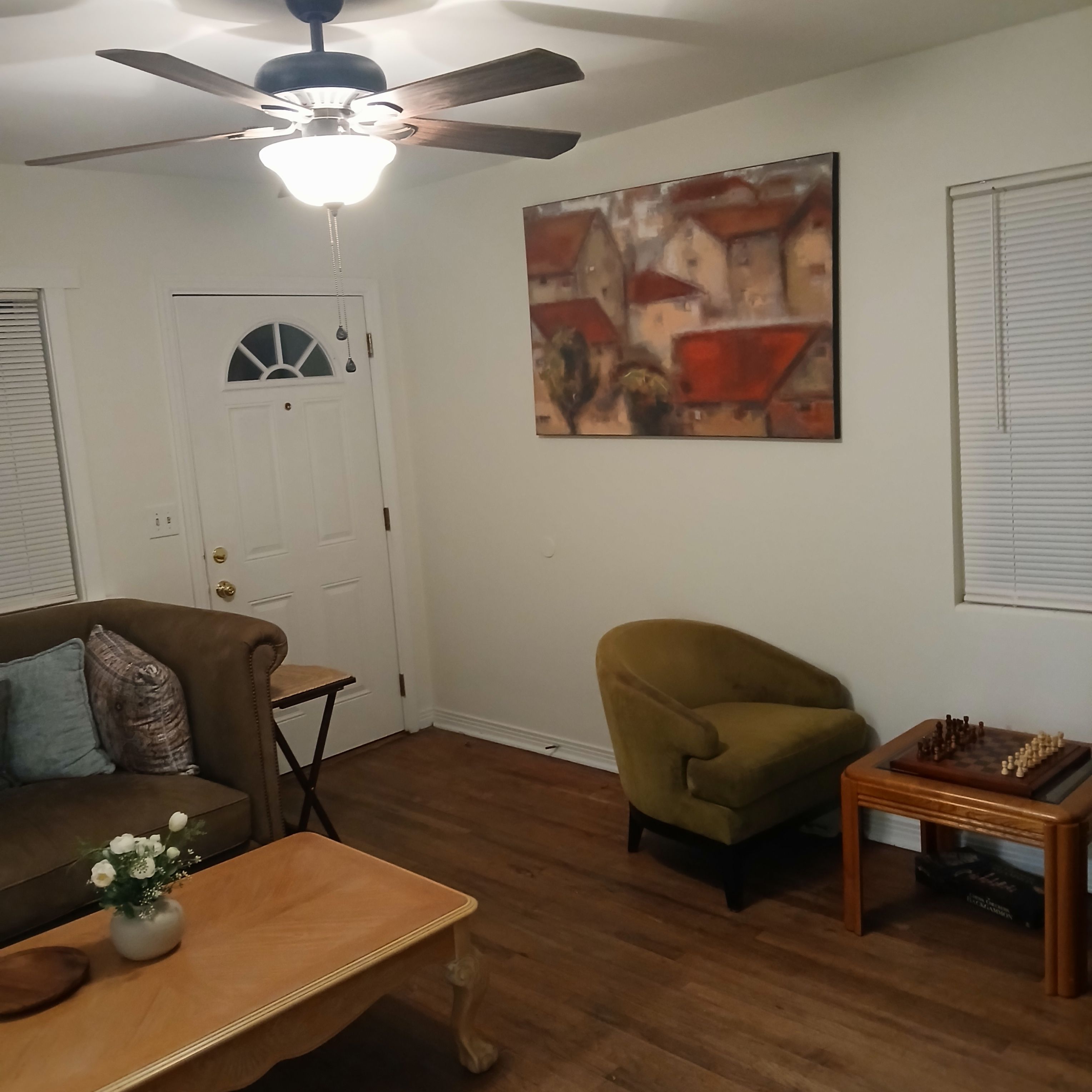 Un salon avec un ventilateur de plafond, une porte avec une fenêtre, un canapé marron, une petite table avec un vase, un fauteuil vert et un jeu d’échecs.