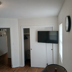 Habitación con televisor de pantalla plana montado en una puerta blanca, armario con ropa colgada y ventana con persianas.