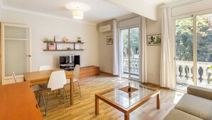 Ein modernes Wohnzimmer mit Glastisch, einem Essbereich mit Holztisch und Stühlen, einem Fernseher und einem Balkon mit Blick auf Bäume.