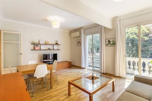 Ein modernes Wohnzimmer mit Glastisch, einem Essbereich mit Holztisch und Stühlen, einem Fernseher und einem Balkon mit Blick auf Bäume.