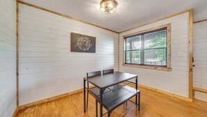 Um quarto com paredes brancas e painéis, piso de madeira, uma mesa com cadeiras, uma janela com persianas e um lustre.